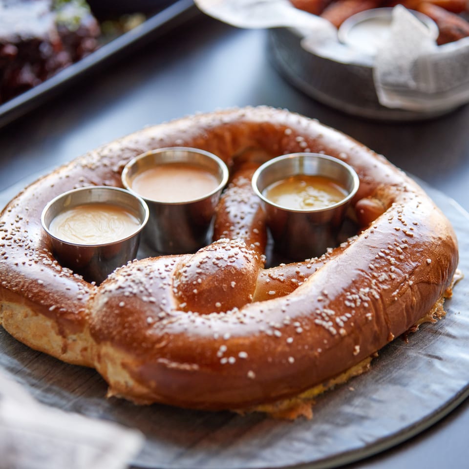 Sesame-topped soft pretzel arranged around three small cups of dipping sauces on a metal serving platter.