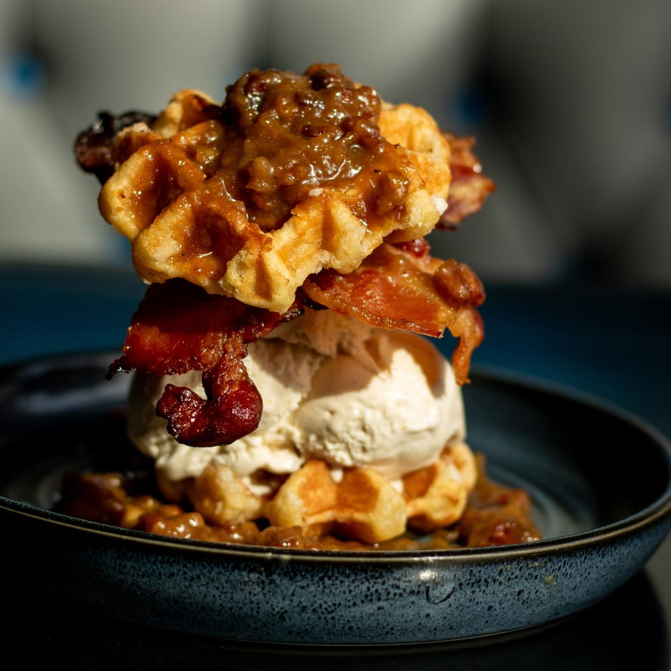 Waffle dessert topped with vanilla ice cream, crispy bacon, and glossy caramelized sauce in a dark bowl.