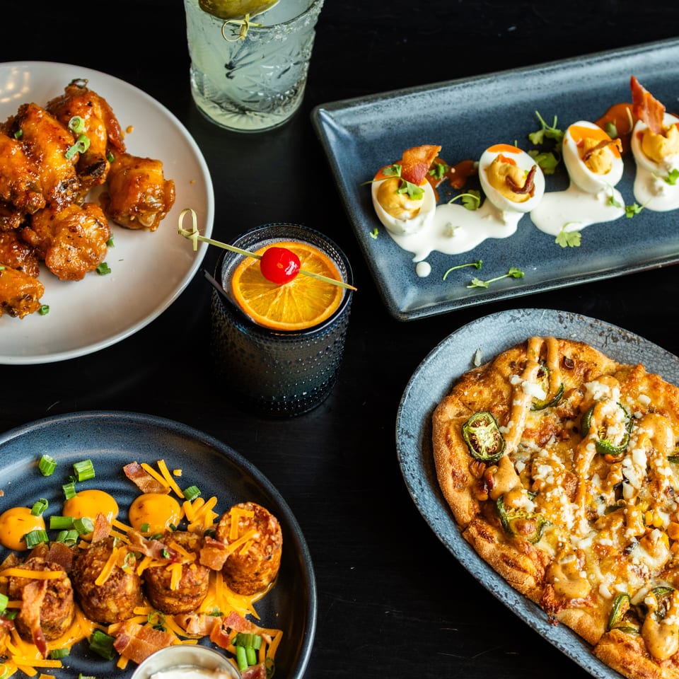 Restaurant spread featuring saucy chicken wings, bacon-topped deviled eggs, loaded flatbread, cheese fries, and colorful cocktails.