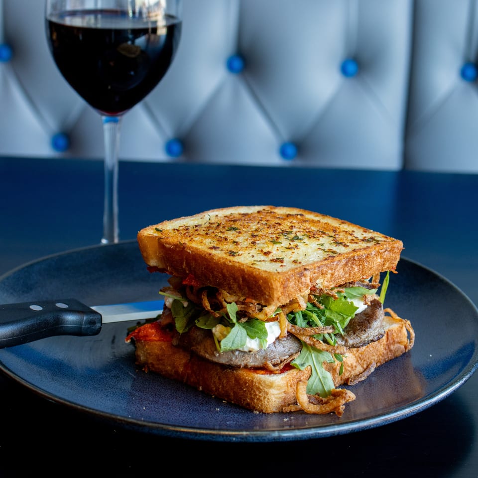 Loaded toasted sandwich with greens, crispy onions, and meat on a blue plate beside a glass of red wine.