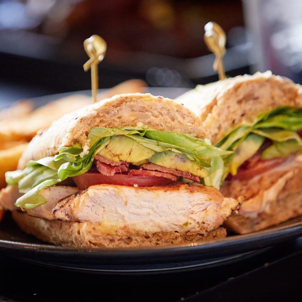 Grilled chicken sandwiches with lettuce, tomato, avocado, and bacon on a black serving plate.