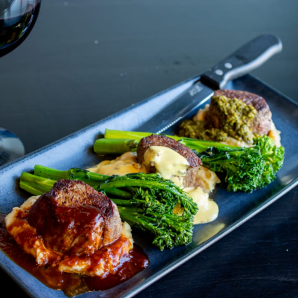 Plated beef medallions with mashed potatoes, broccolini, and creamy sauce on a rectangular dish beside a glass of red wine.