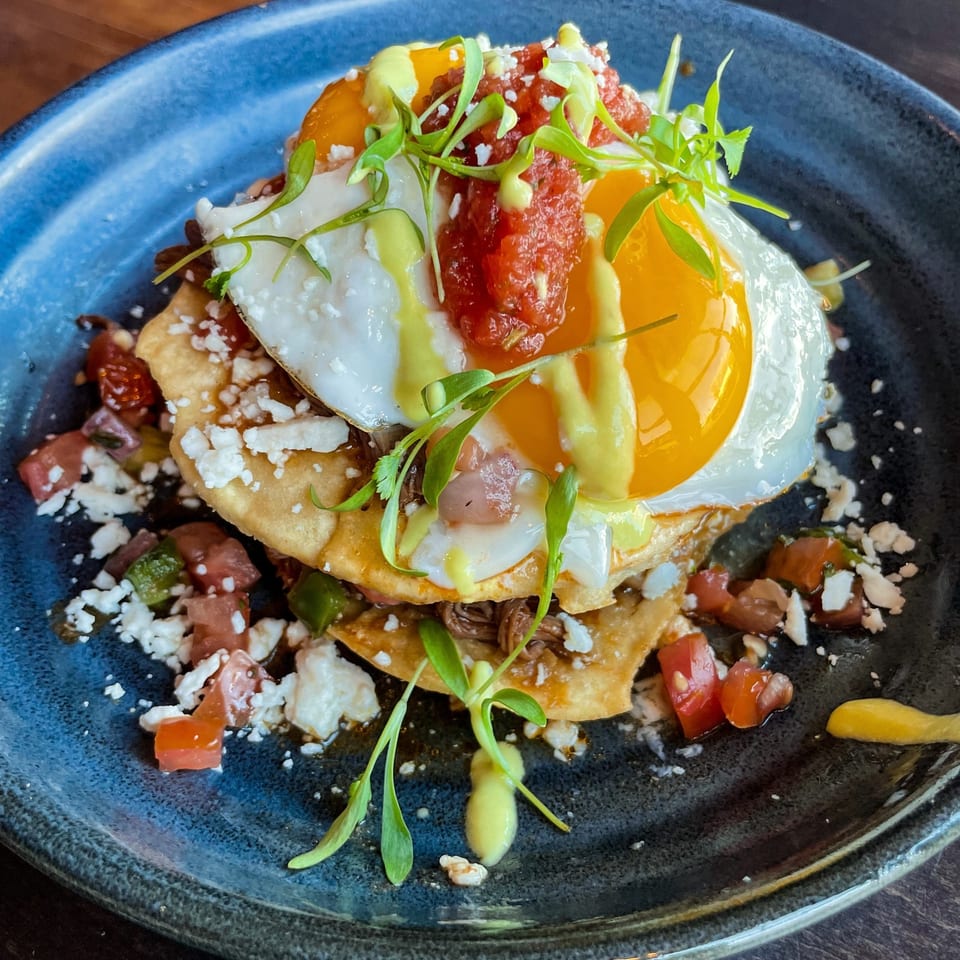 Stacked crispy tortillas topped with sunny-side-up eggs, chorizo, avocado sauce, queso fresco, pico de gallo, and fresh herbs on a blue plate.