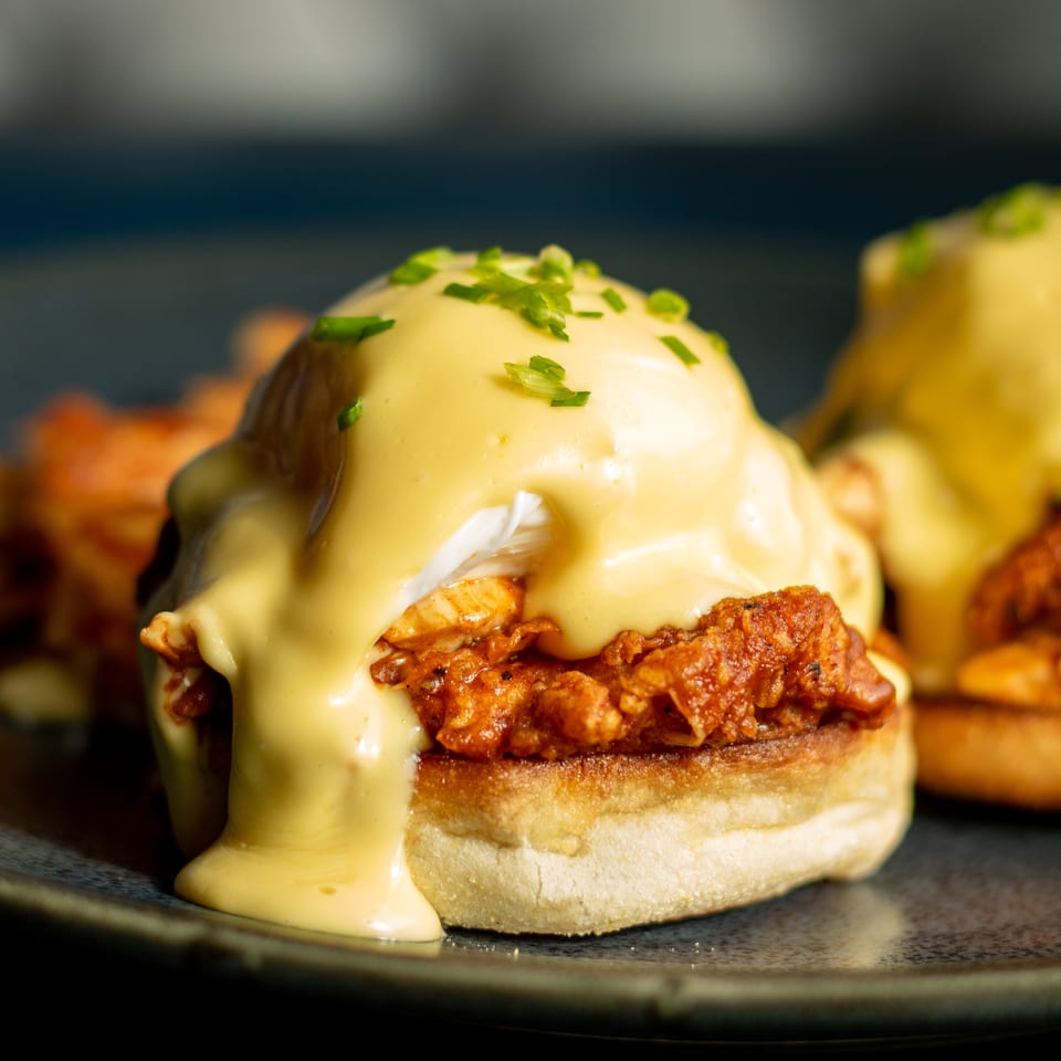 Two crispy chicken sandwiches on buns topped with creamy hollandaise sauce and chopped green onions on a dark plate.