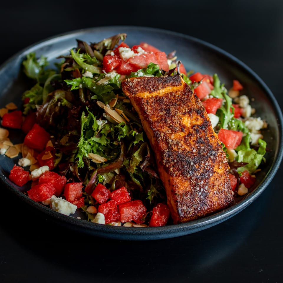 Grilled salmon fillet served over mixed greens with diced tomatoes, crumbled feta, and sliced almonds in a dark bowl.