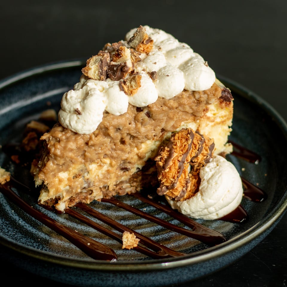 Slice of layered cake with creamy frosting, chocolate drizzle, whipped cream, and crunchy chocolate-covered topping on a dark plate.