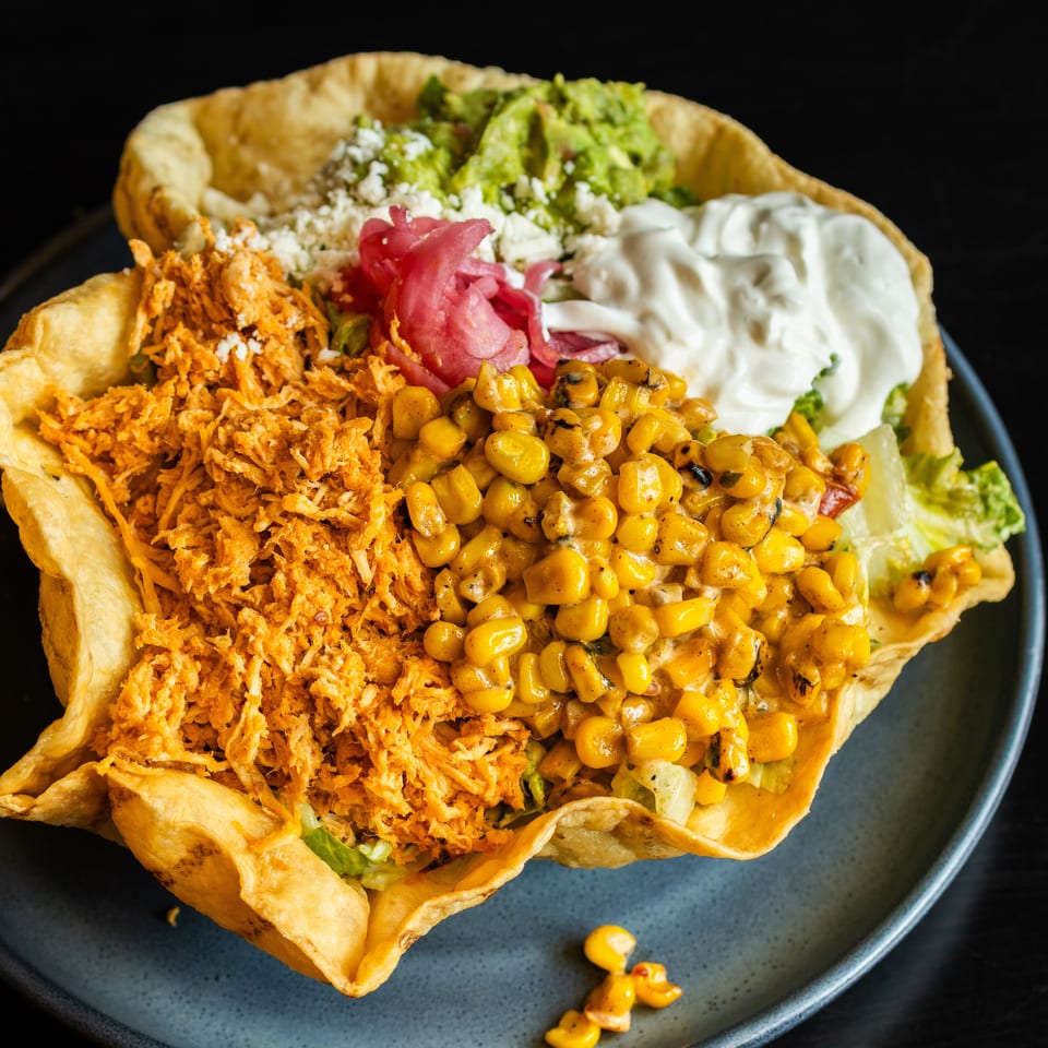 Crispy tortilla bowl filled with shredded chicken, grilled corn, lettuce, guacamole, pickled onions, feta, and sour cream.